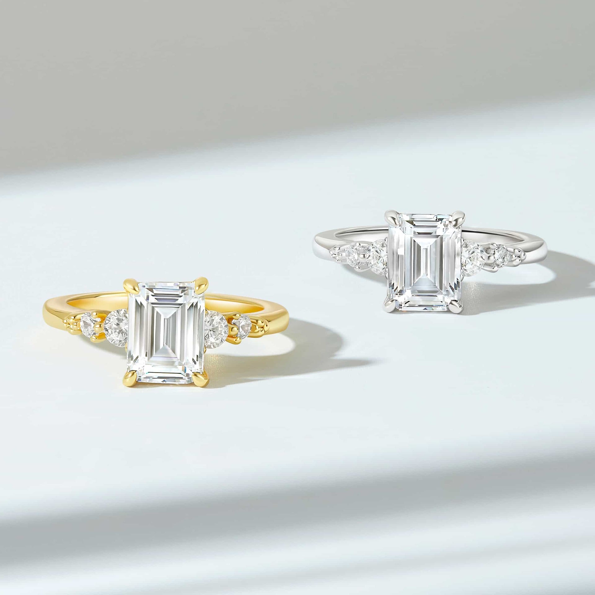 a modern 1.5 carat emerald cut engagement ring with two stunning round cut side stones on both sides shown in gold and silver