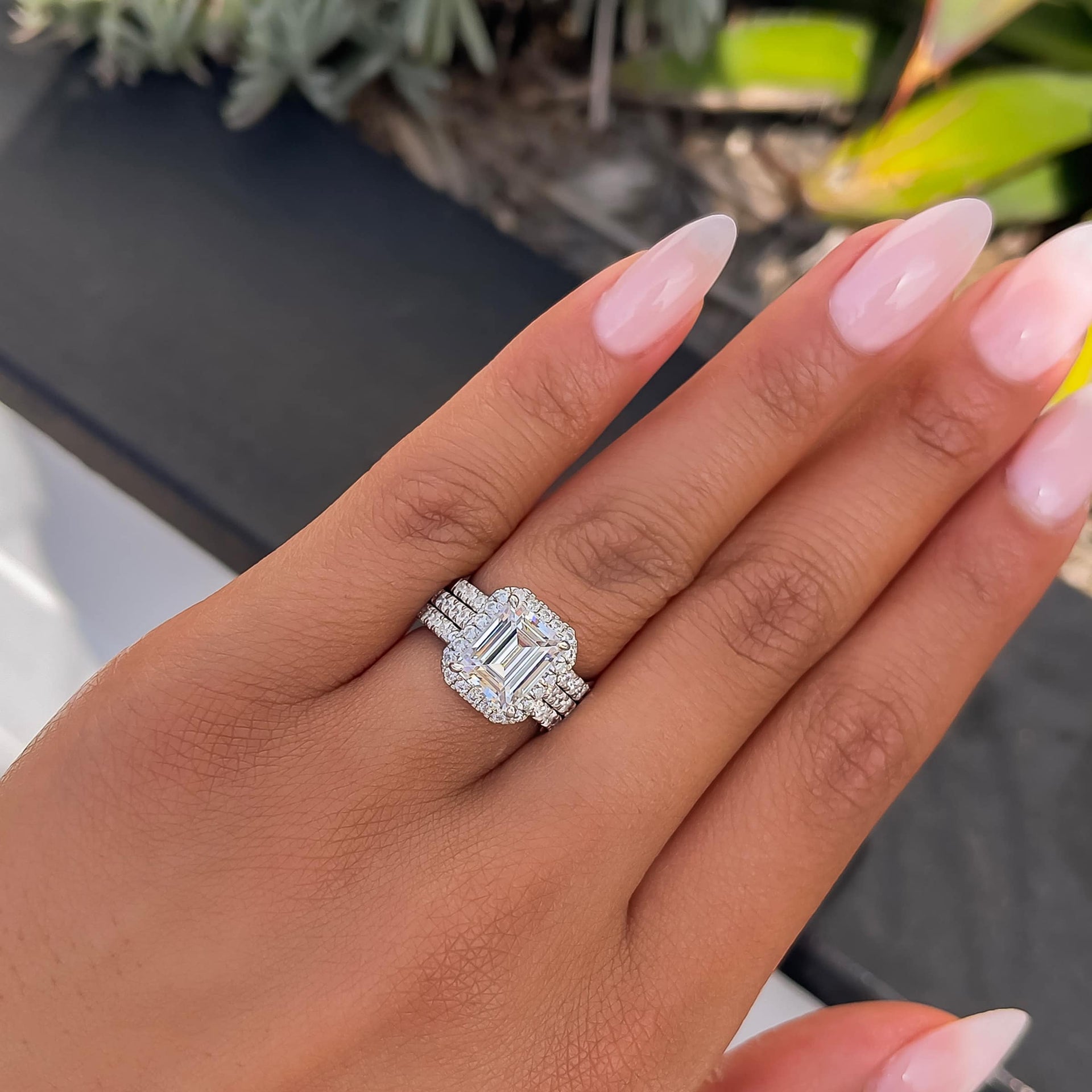 stunning emerald cut engagement ring with halo and half eternity band paired with two silver wedding bands on model with pink nails