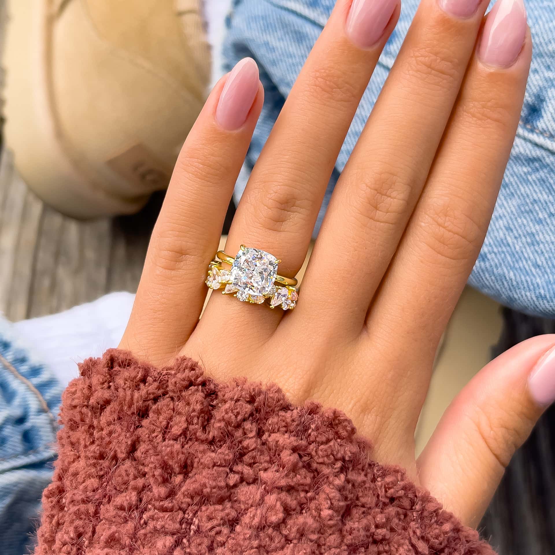 Close-up of The Claire gold engagement ring with 3.5 carat cushion-cut simulated diamond, stacked with gold multi-stone wedding band on hand with pink nails
