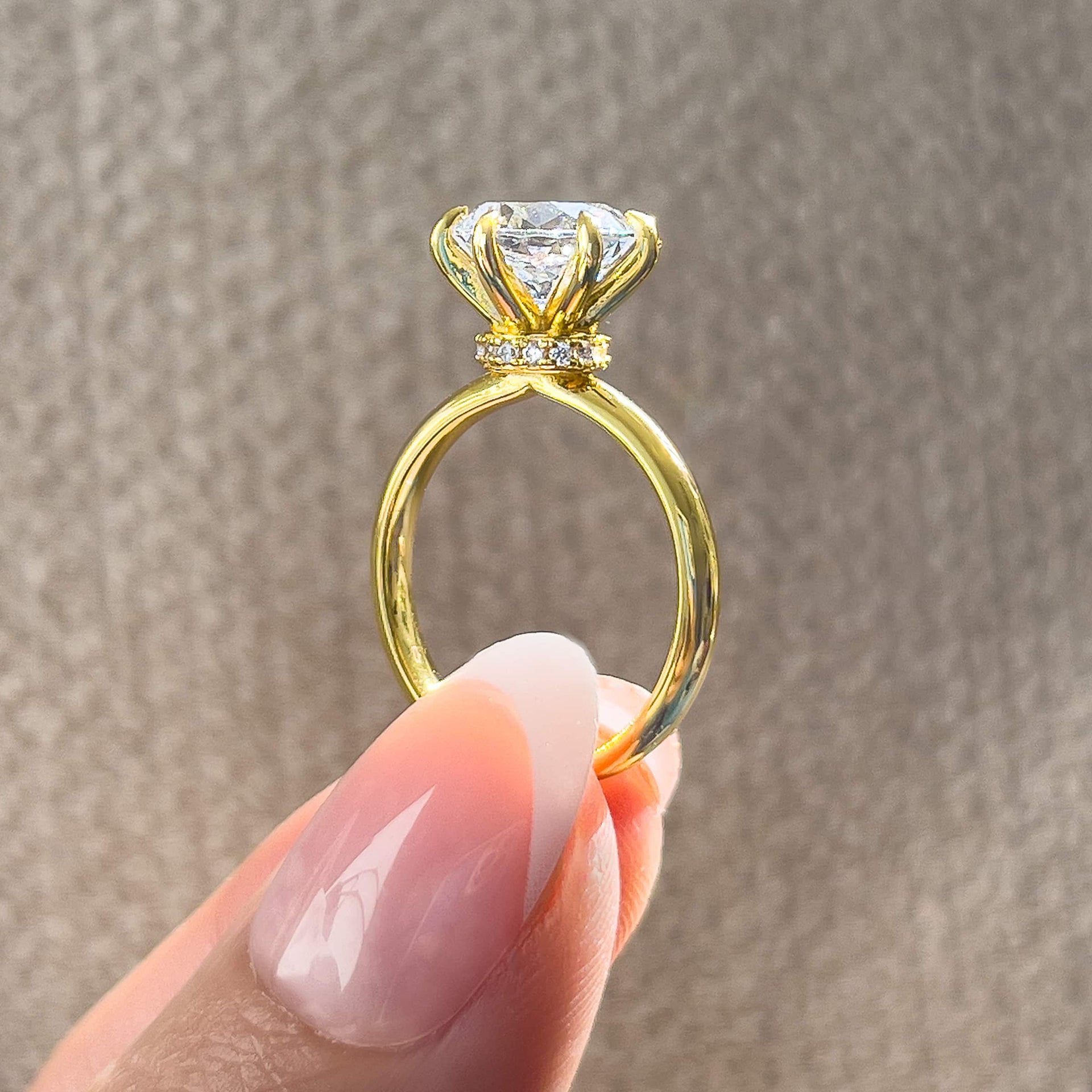 Side view of gold 6 prong 2 carat round cut solitaire engagement ring with secret halo detail held by model with french tip nails