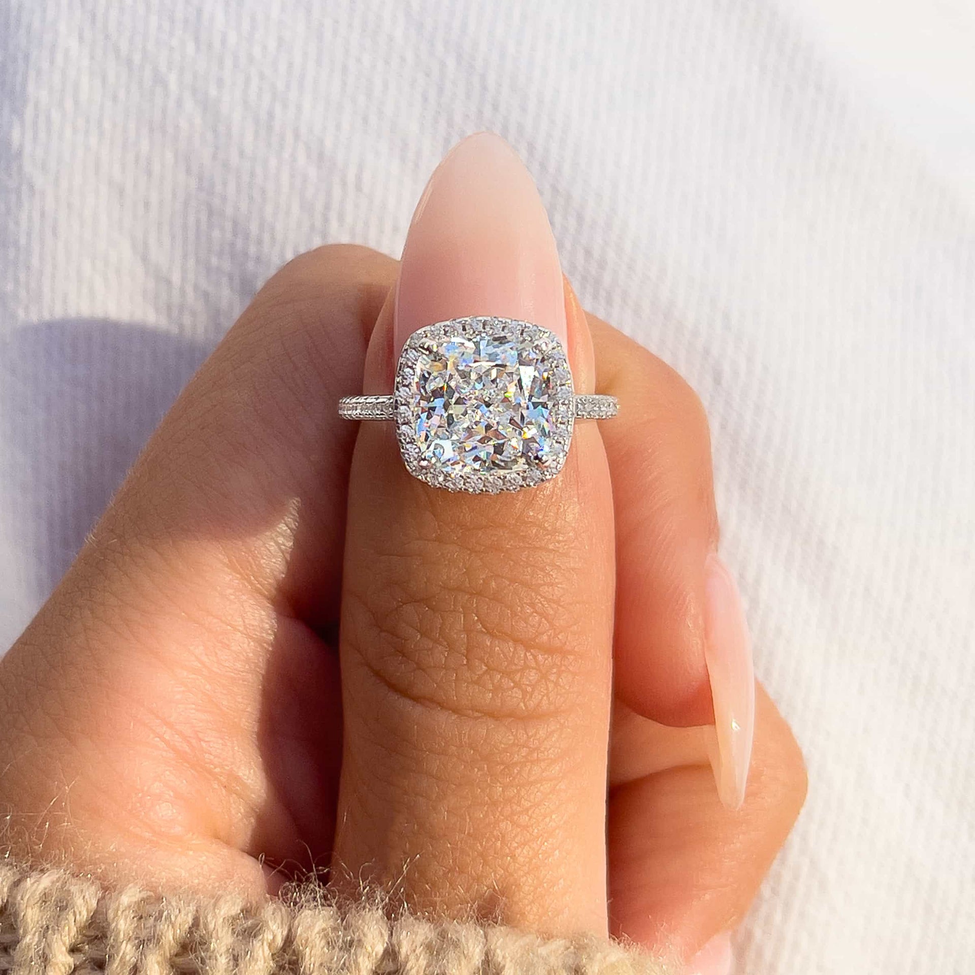 stunning 3 carat silver cushion cut engagement ring on model's thumb with pink manicured nails and white pants in background