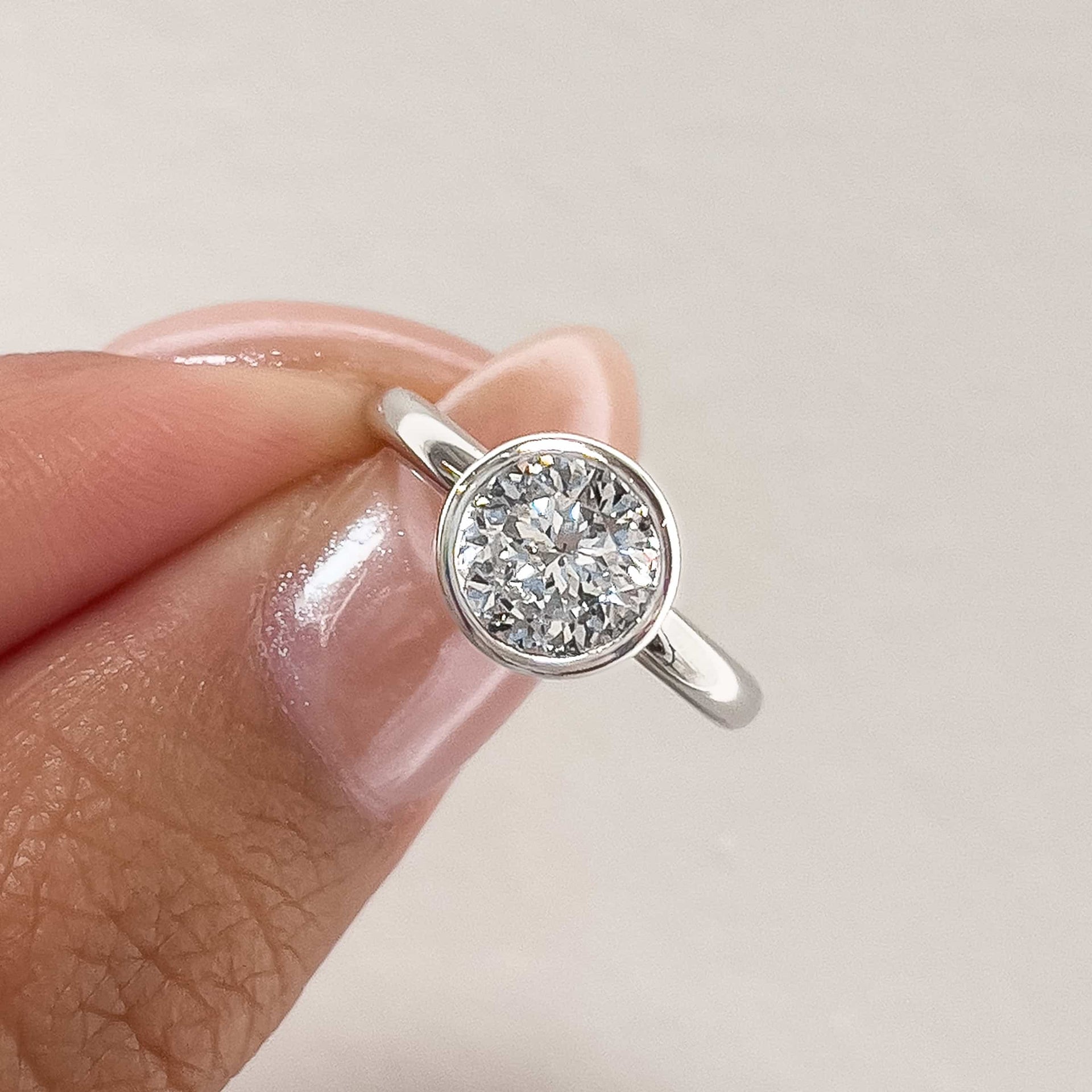 a beautiful silver round cut bezel setting engagement ring pinched between models fingers with pink shimmering nails