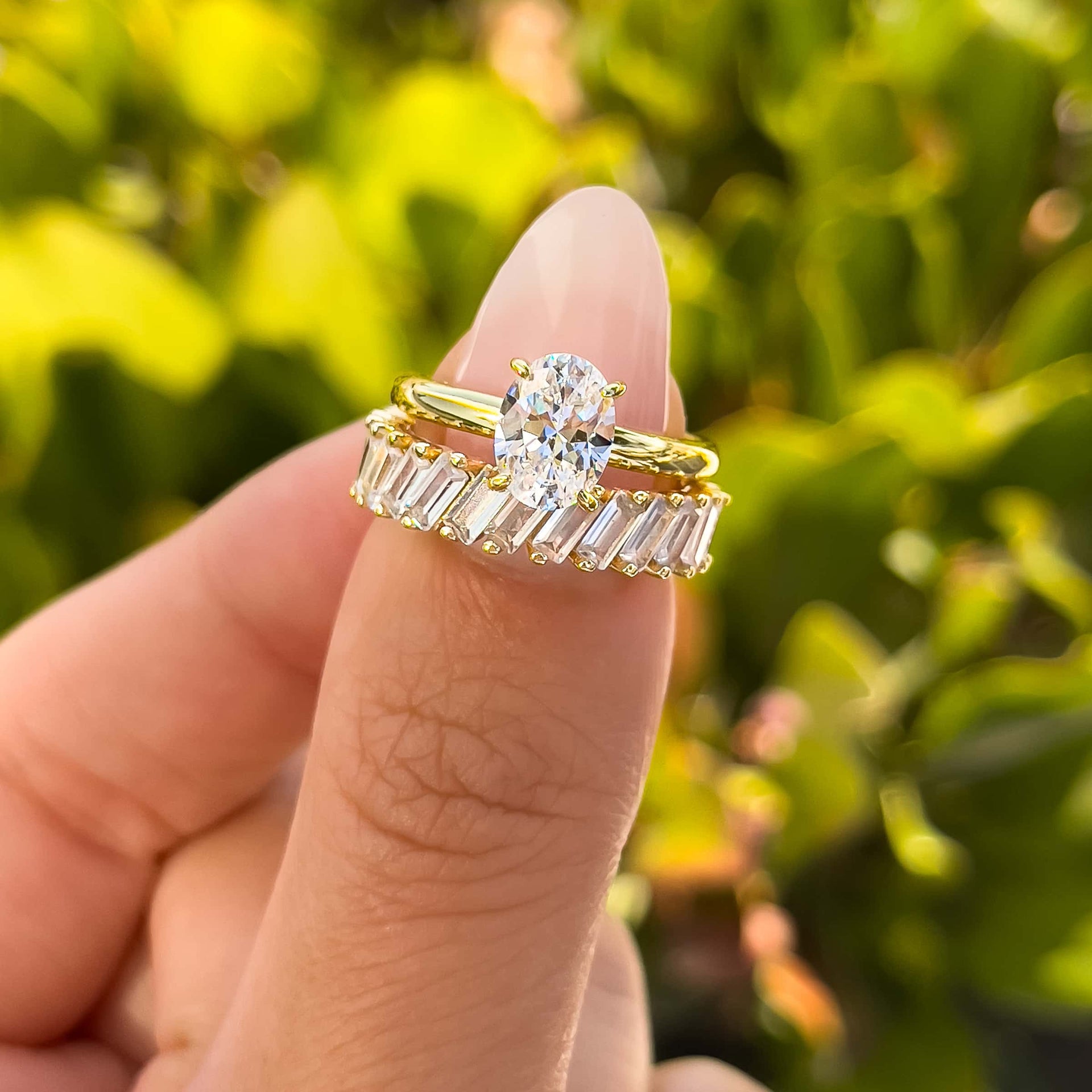 beautiful gold solitaire engagement ring paired with sparkling baguette band with inset stones on model's thumb with greenery in background