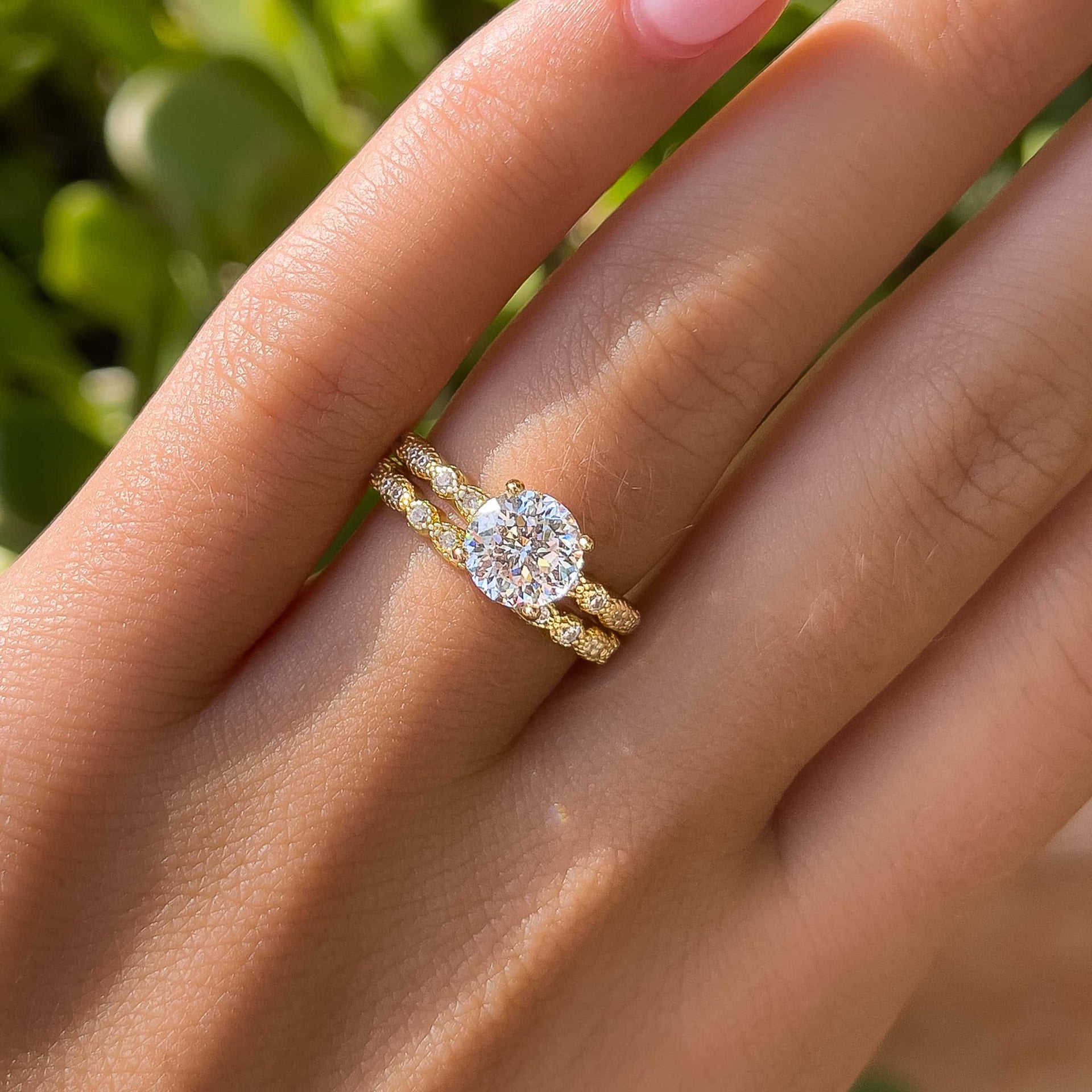 woman wearing gold 1.5ct round cut center stone engagement ring with matching wedding band and greenery in the background