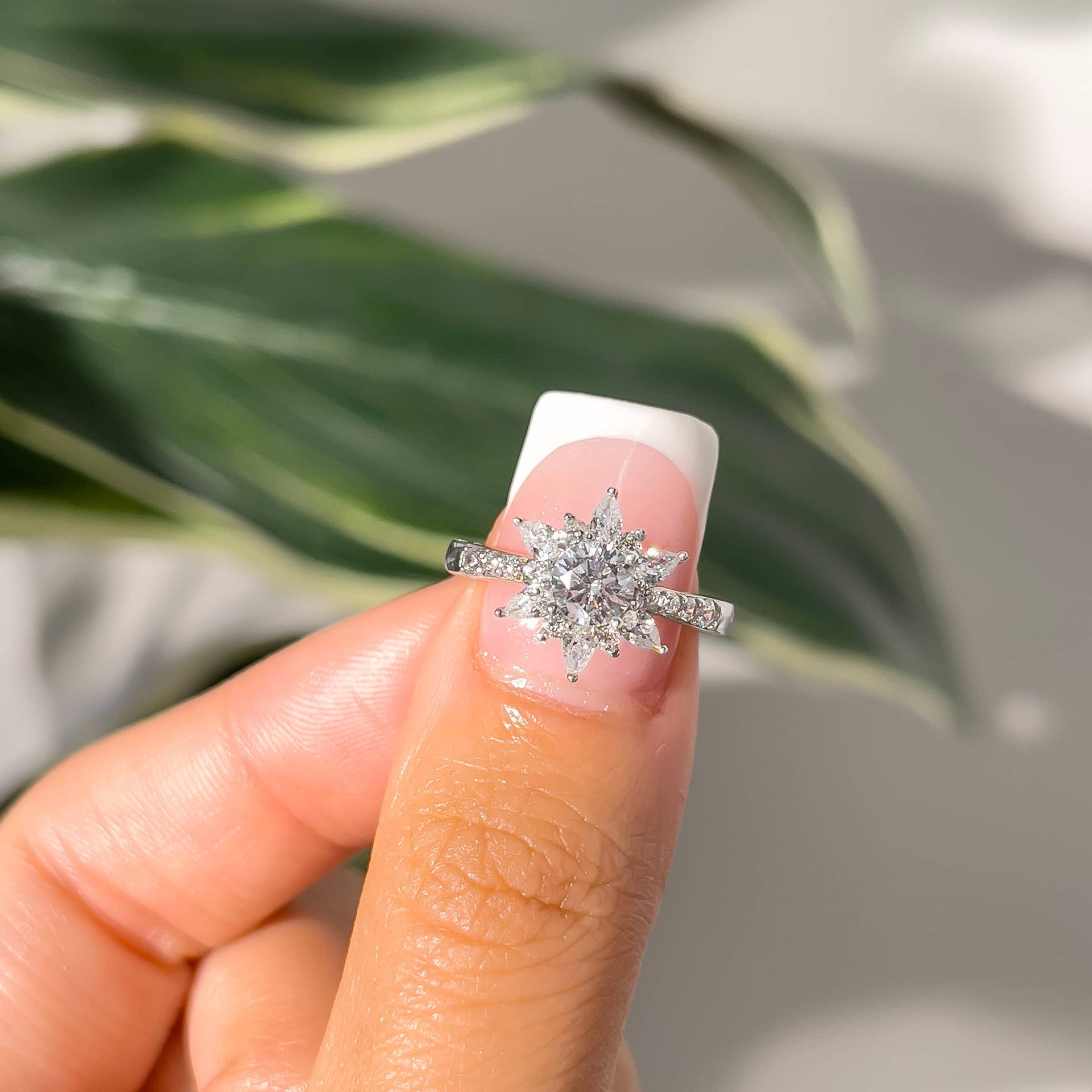 unique silver engagement ring with a round cut center stone and various stones around it making a star-like shape, on thumb with french tip nail